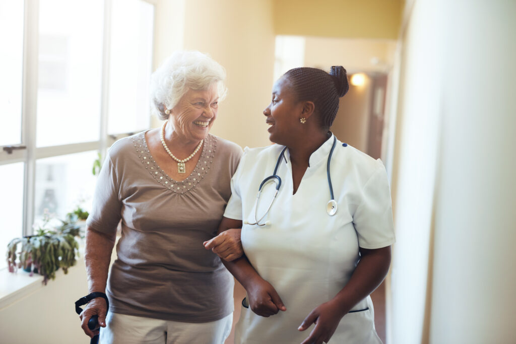 Senior woman walking in the nursing home supported by a caregiver. Nurse assisting senior woman.